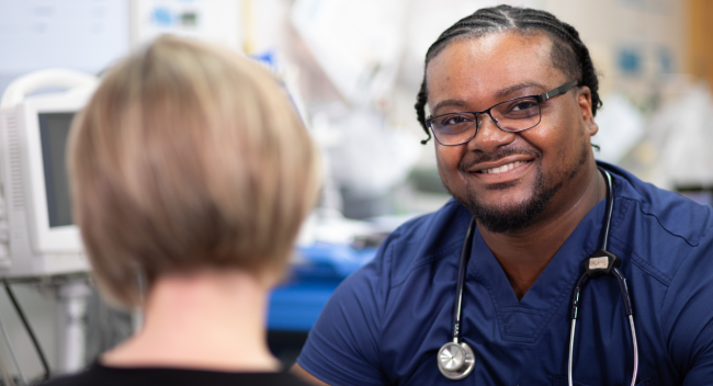 Tristan Lollis is pictured in a hospital exam room wearing blue scrubs. He's smiling toward the camera over the shoulder of a mock patient.